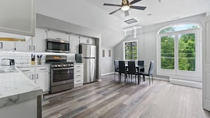 A modern kitchen with stainless steel appliances, a dining area with a table and chairs, and a large window offering a view of greenery.