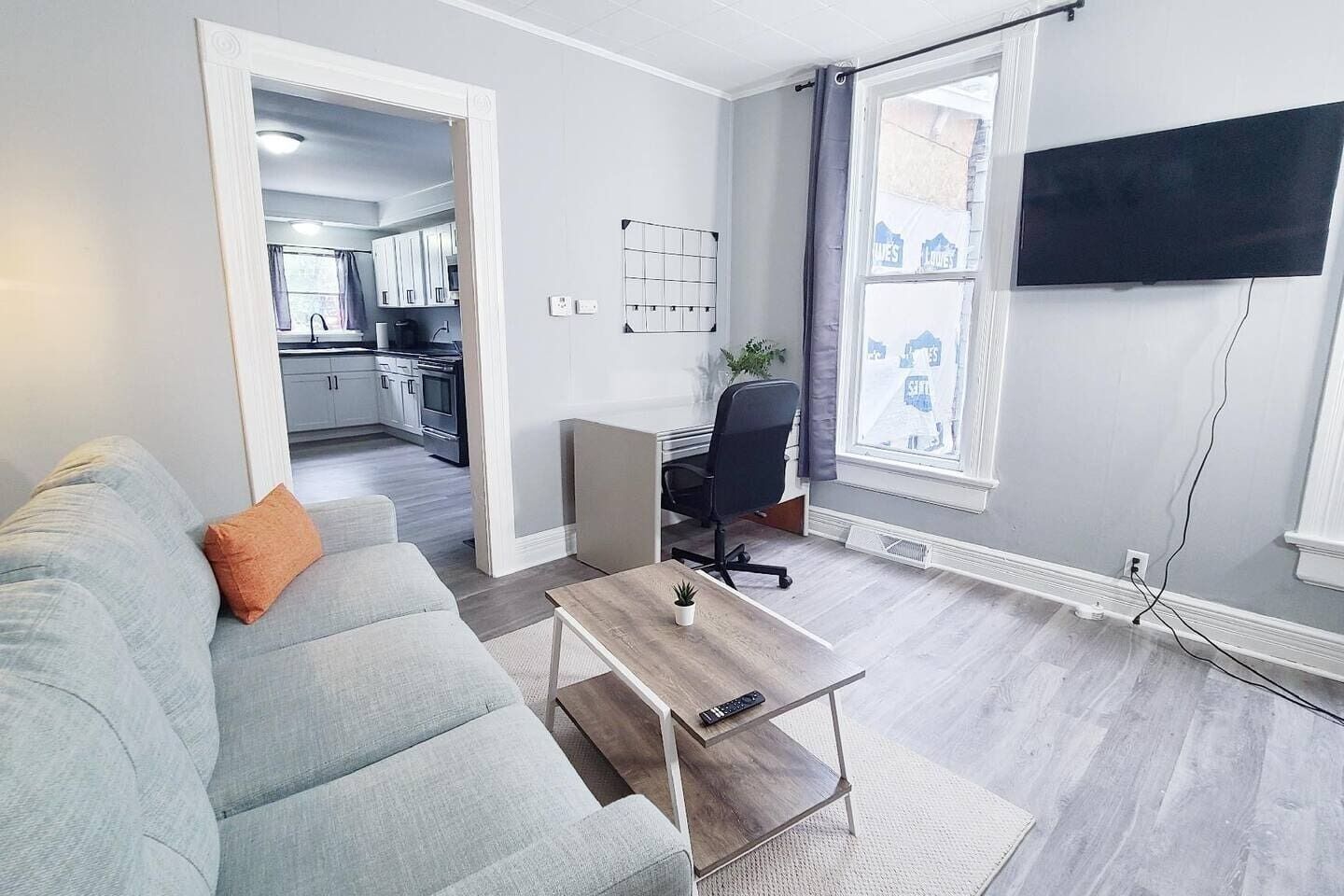 A modern living room with a grey sofa, a coffee table, a TV mounted on the wall, and a kitchen visible through an open door.
