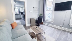 En moderne stue med en grå sofa, et sofabord, et fjernsyn ophængt på væggen, og et køkken der kan ses gennem en åben dør.