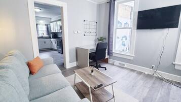 A modern living room with a grey sofa, a coffee table, a TV mounted on the wall, and a kitchen visible through an open door.
