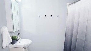 Et badeværelse med en hvid vask, spejl og skab, bruseforhæng og en hvid måtte på gulvet.