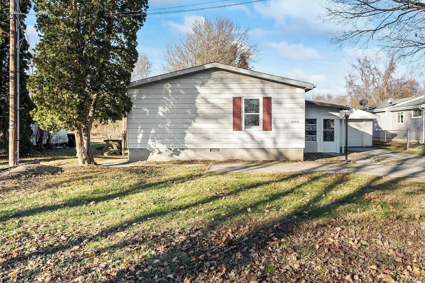 A single-story mobile home with a garage, a small attached shed, and a tree in the foreground.