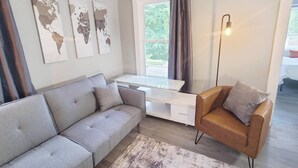 Un salon moderne avec un canapé gris, un fauteuil marron, un bureau blanc et une grande fenêtre avec des rideaux.