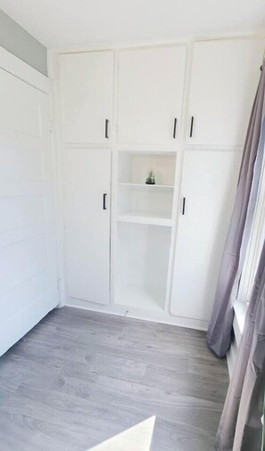 A room with a white wardrobe, a small shelf, and a grey wooden floor.
