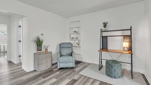 A modern living room with a grey armchair, a wooden console table with plants, a metal shelving unit with a lamp, and a grey ottoman.