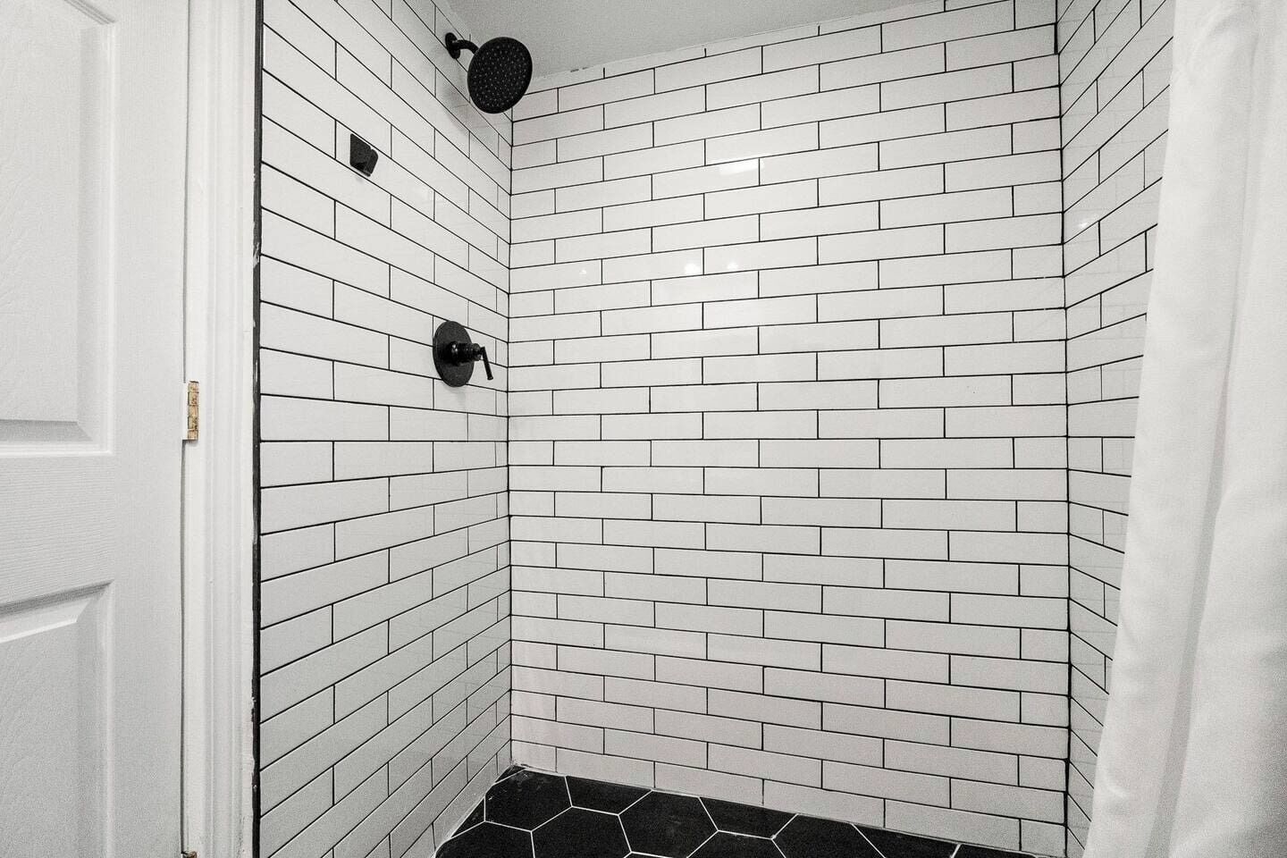 A bathroom with white subway tiles, a shower with a black head, and a black hexagonal tile floor.