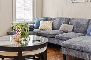 Un canapé modulable agrémenté de plusieurs coussins, une table basse en verre ornée d’un objet décoratif et une fenêtre avec des stores.