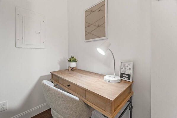 A wooden desk with a white lamp, a potted plant, and a framed sign on a white wall.