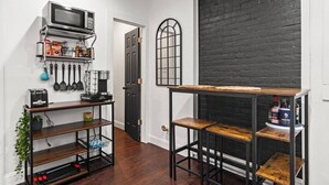 Une cuisine équipée d’un four à micro-ondes, d’une machine à café et de divers ustensiles de cuisine. On y trouve une table en bois avec des tabourets, ainsi qu’un mur en briques noires.