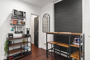 A kitchen with a microwave, coffee maker, and various kitchen utensils. There is a wooden table with stools and a black brick wall. - Classic Brick Apartment | Convenient Parking (Fort Wayne)