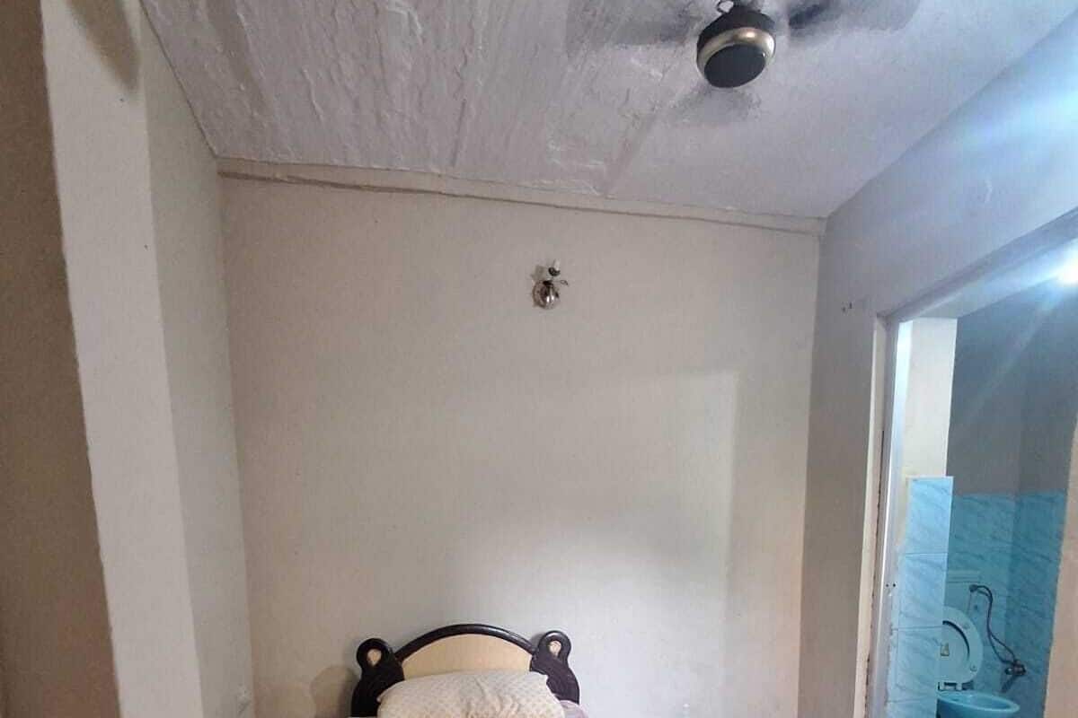 Una habitación pequeña con una cama individual, una colcha roja, un ventilador de techo y una pared blanca lisa.