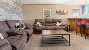 A living room with two brown leather sofas, a wooden coffee table, and a kitchen area with a window, artwork, and bar stools. - Mountain Farm Stay, Alpacas & Views, Royal Gorge 15min (Cañon City)