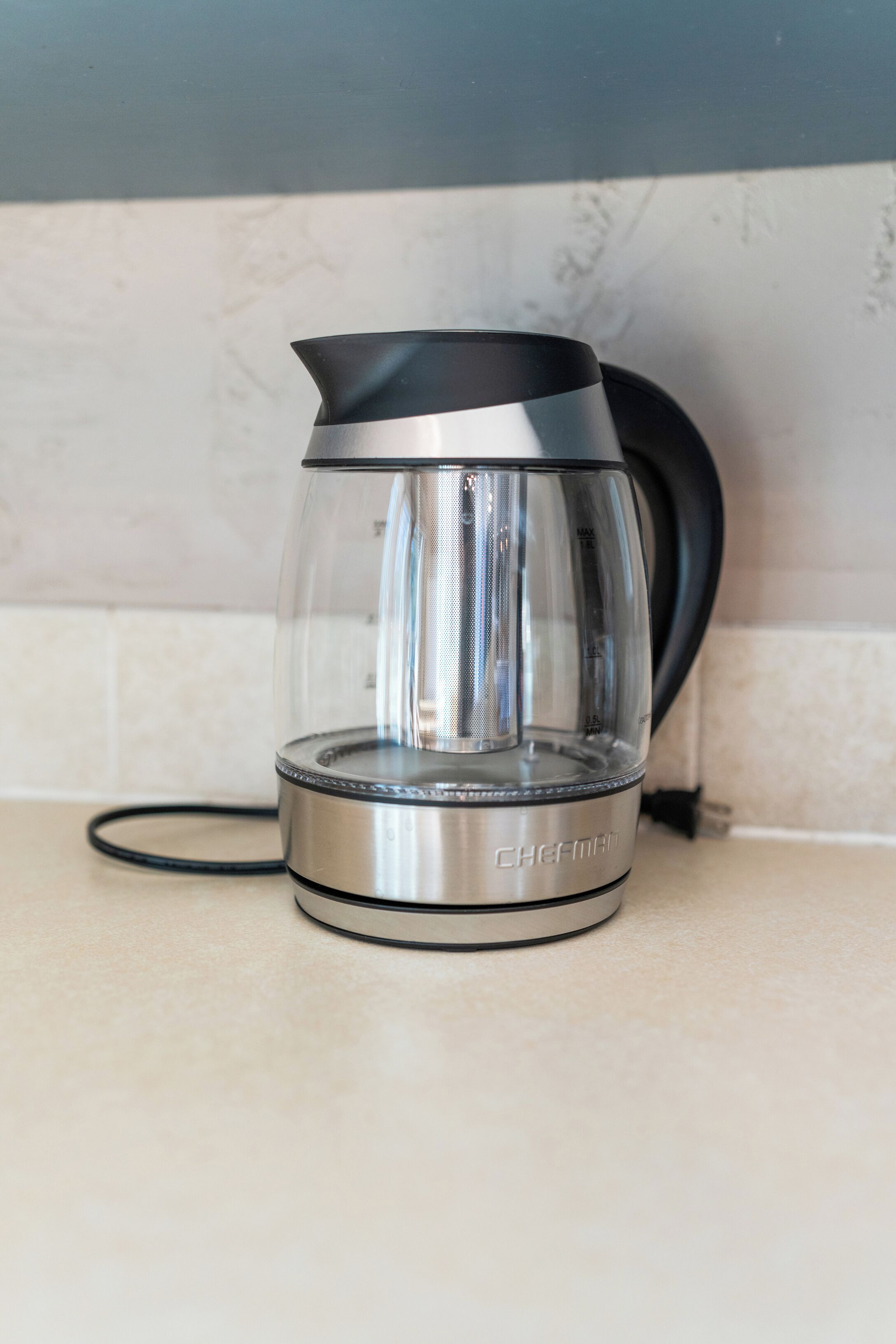 A stainless steel electric kettle with a transparent glass carafe, placed on a kitchen counter.