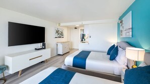 Une chambre d’hôtel moderne dotée d’un grand téléviseur à écran plat, d’une console blanche, de deux lits aux touches de couleur bleue, d’un mur d’accent de couleur turquoise et d’une petite kitchenette.