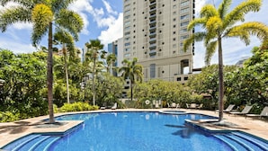 Una piscina rodeada de palmeras y sillones de descanso, con un edificio alto al fondo.