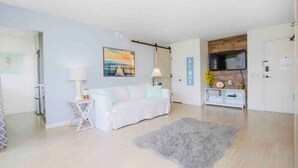 Un salon moderne avec un canapé blanc, un tapis gris, un téléviseur fixé à un mur en bois et une petite table d’appoint avec une lampe.