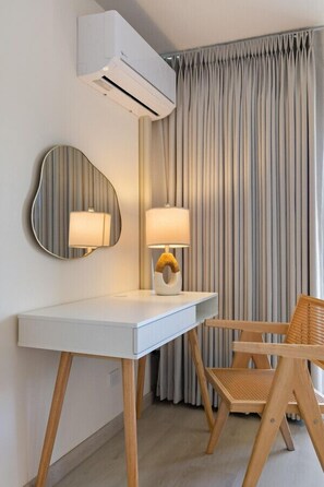 Une chambre d’hôtel moderne avec un bureau blanc, un miroir rond, une lampe, un fauteuil en osier et une grande fenêtre avec des rideaux.