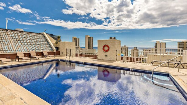 Une piscine sur le toit, avec une eau d’un bleu cristallin, entourée de chaises longues beiges et agrémentée d’une bouée de sauvetage fixée à un mur blanc.