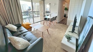 Un salon moderne avec un canapé, une table à manger et des chaises. On y trouve un téléviseur posé sur un meuble et une fenêtre avec des rideaux.