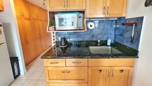 A kitchen with wooden cabinets, a black countertop, a microwave, a kettle, and a sink.
