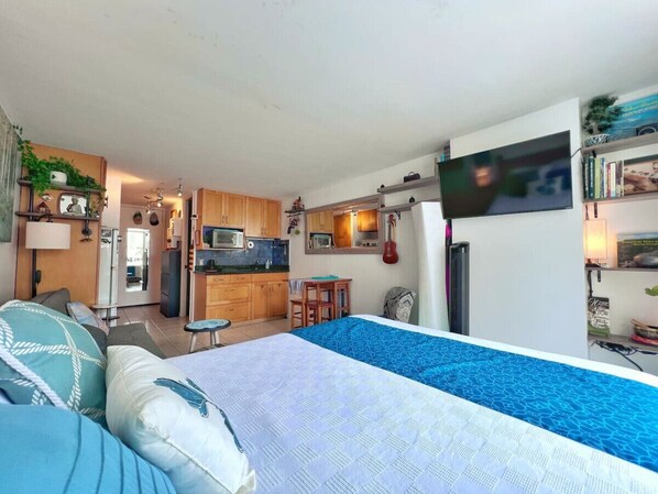 A compact studio apartment with a kitchenette, a bed with a blue patterned bedspread, a TV mounted on the wall, and a small dining table. - Waikiki Beach Studio: Walk 2 Surf + Assigned Prkng (Honolulu)