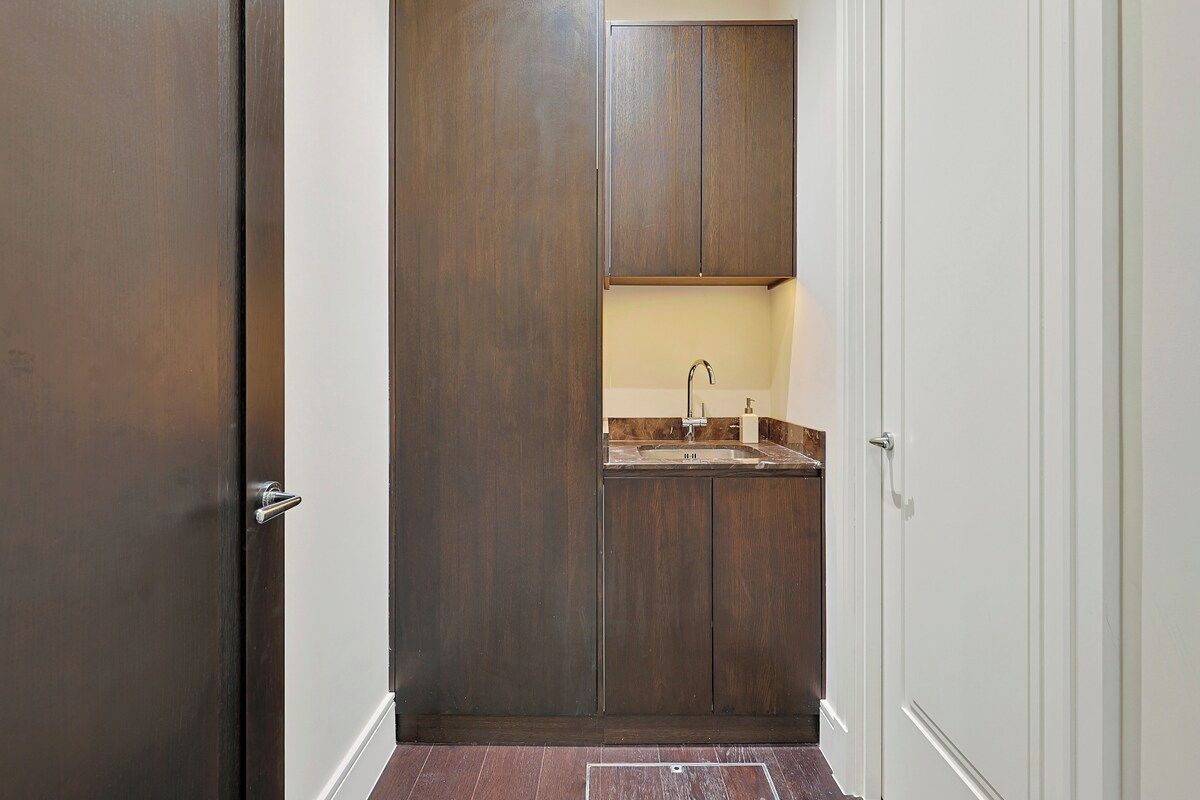 A narrow hallway with a wooden door on the left, a white door on the right, and a small kitchenette area with a sink and countertop.