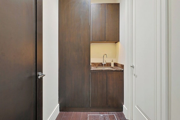 A narrow hallway with a wooden door on the left, a white door on the right, and a small kitchenette area with a sink and countertop.
