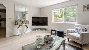 Ein modernes Wohnzimmer mit Fernseher, rundem Couchtisch mit Dekoration, einer Couch und einem großen Fenster mit Blick ins Freie.