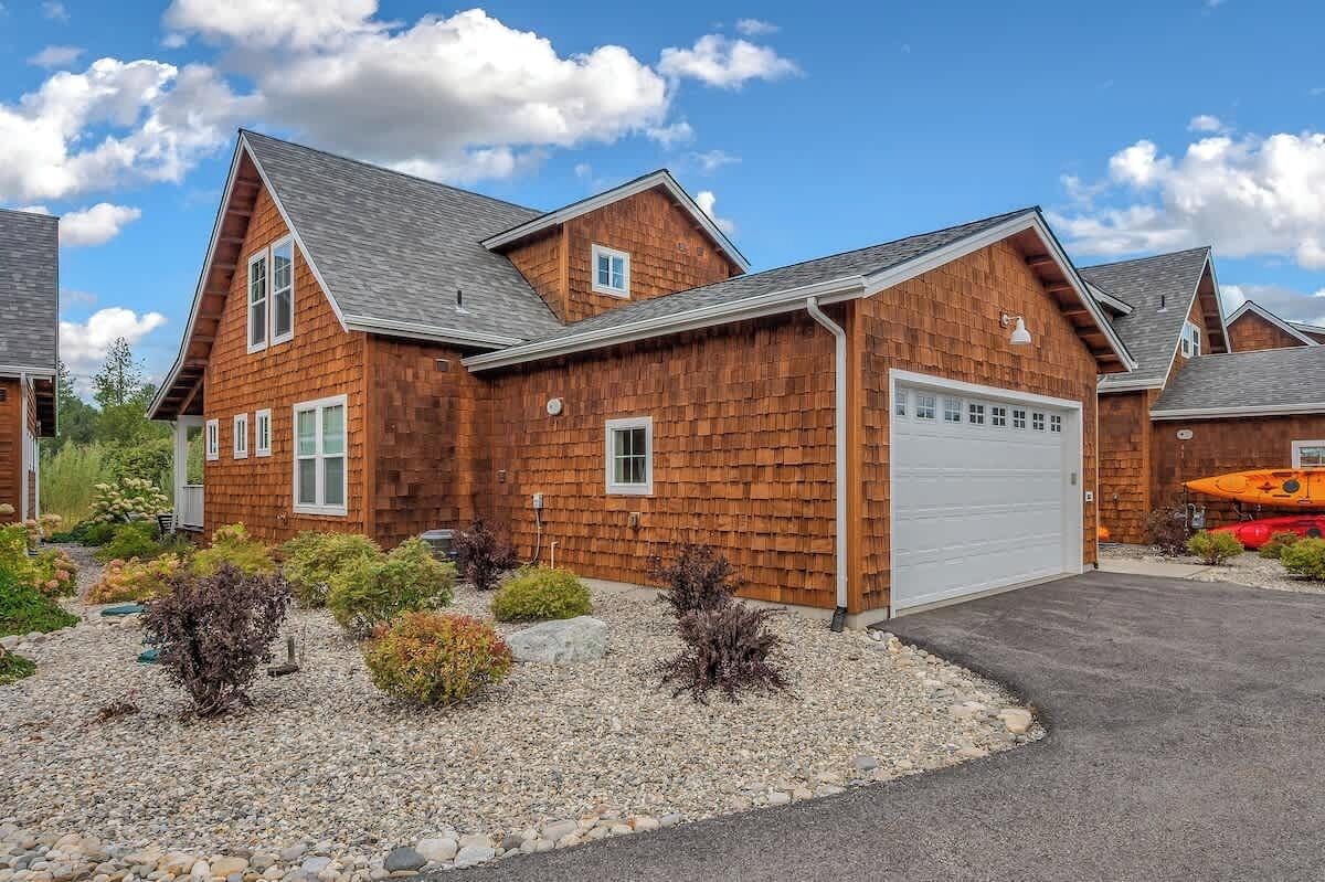 A two-story wooden house with a garage, surrounded by a gravel driveway and small shrubs.