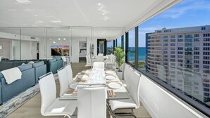 A modern dining area with a long table set for a meal, large windows offering a view of the ocean and a high-rise building, and a seating area with sofas and a TV screen. - Luxury Ocean View Condo / Beach Service / 1521 (Miami Beach)