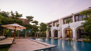 A swimming pool with lounge chairs and umbrellas, surrounded by greenery and a modern building with arched windows. - Stone Age Boutique  (Kampot)