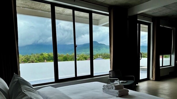 A hotel room with a large bed, a chair, and a view of a body of water and mountains. - Stone Age Boutique  (Kampot)