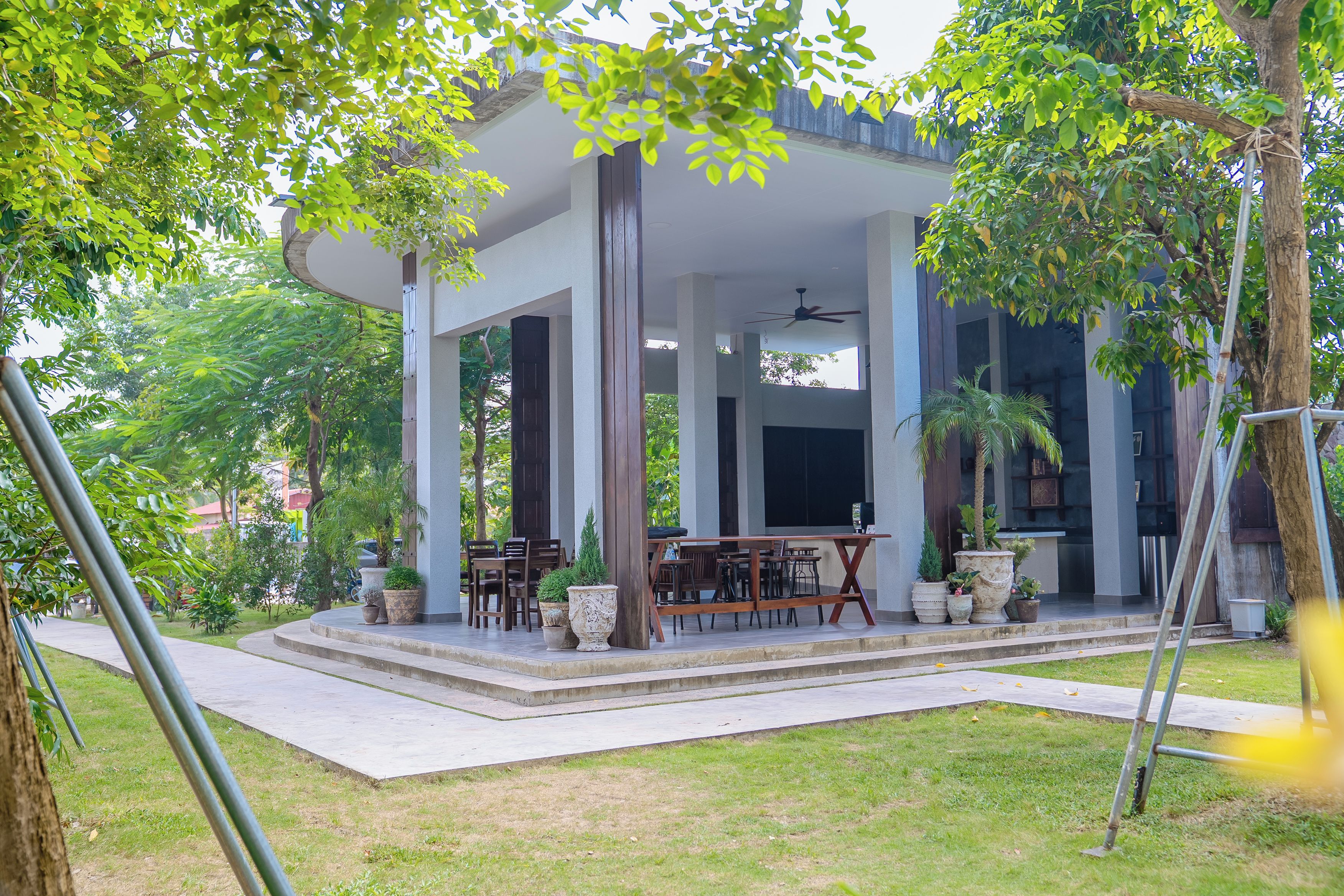 A covered outdoor seating area with tables and chairs, surrounded by greenery and trees.