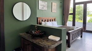A room with a green wall, a round mirror, a wooden headboard bed, a small bedside table with a bowl, and a window with curtains. - Stone Age Boutique  (Kampot)