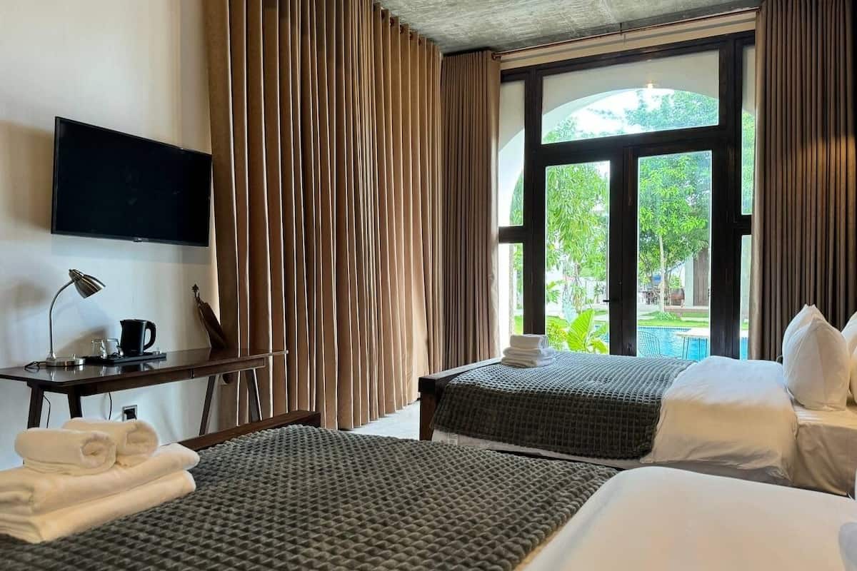 Una camera d'albergo con due letti, una scrivania con una lampada, una televisione e una finestra con tende.