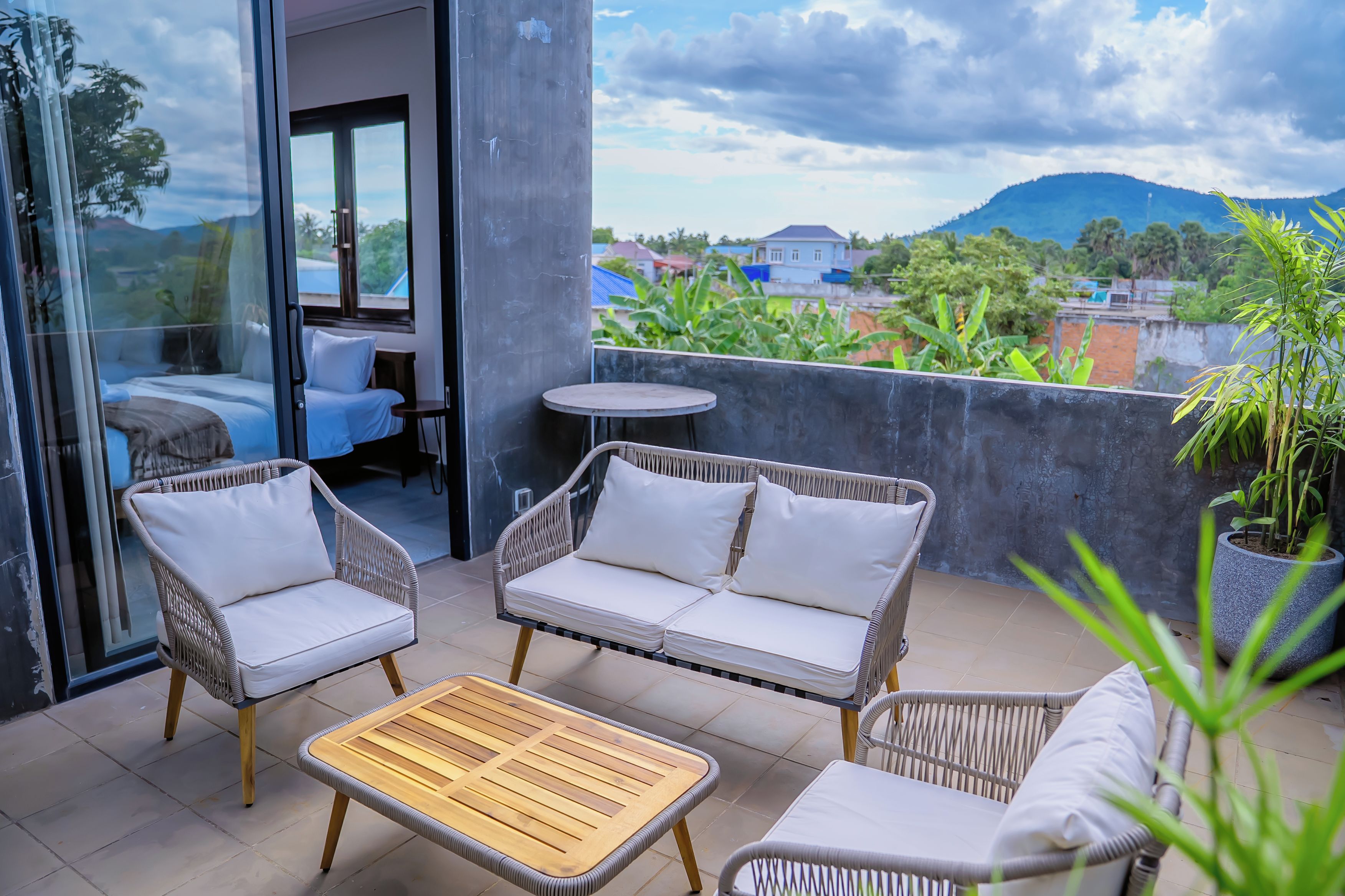 Un balcón con mobiliario de mimbre, una mesa de madera y vistas a una zona residencial con casas y vegetación.