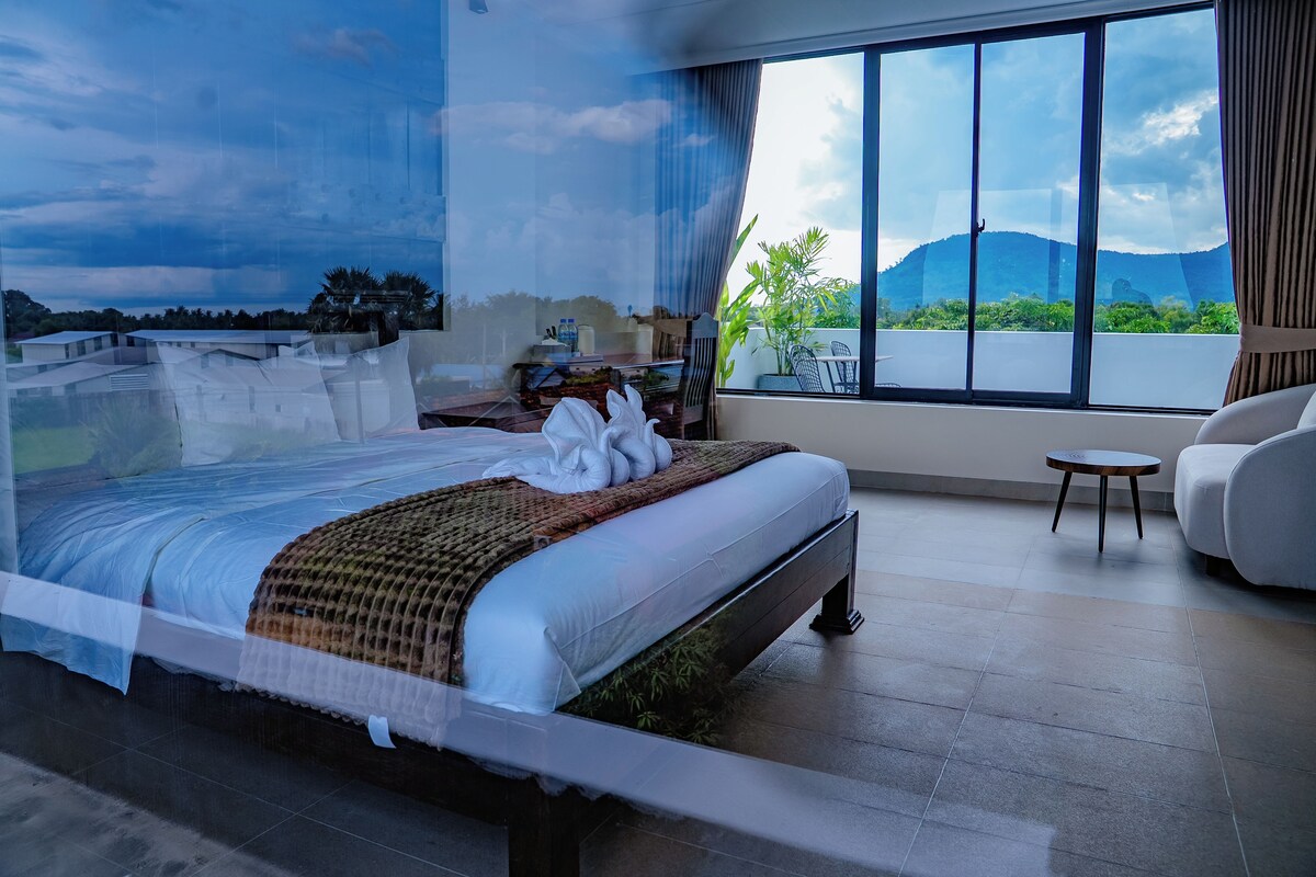 A bedroom with a large bed, a view of the outdoors through a window, and a small table.