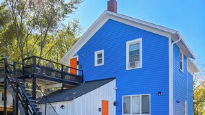 Casa de dos pisos con fachada azul, puerta blanca y escalera negra que lleva a un balcón.