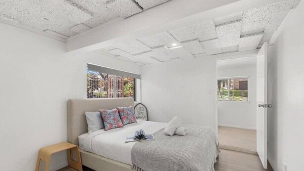 Ein modernes Schlafzimmer mit einem Bett, einem kleinen Holzschemel, einem Fenster mit Blick auf Gebäude und einer Tür zu einem anderen Raum.