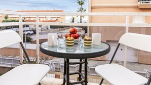 A balcony with a round table set for two, featuring a glass top, white chairs, and a fruit bowl with apples and pastries. - Llevant by Interhome (Creixell)