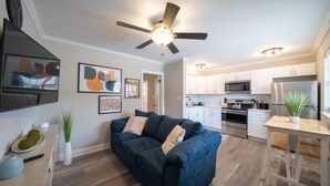 A modern living room with a dark blue sofa, a ceiling fan, and a kitchen area with white cabinets and stainless steel appliances. - OV Mosaic - Coconut Suite (Norfolk)
