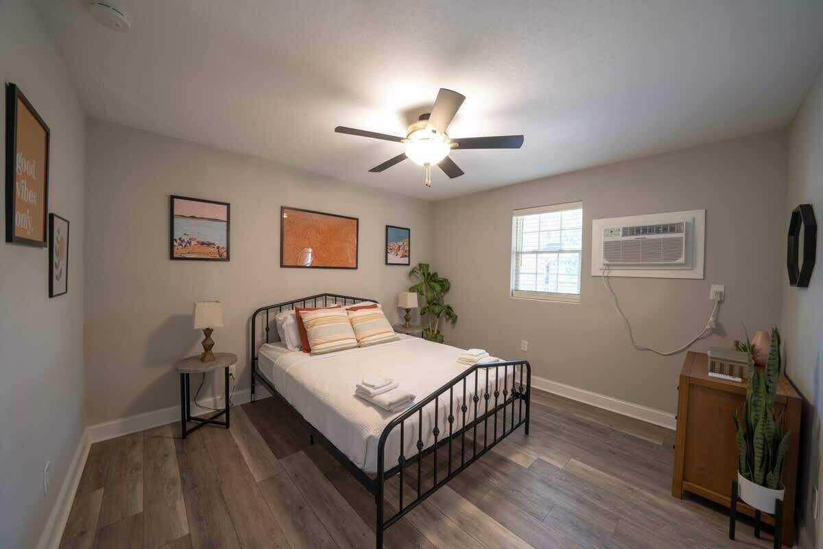 A bedroom with a bed, a ceiling fan, a window with an air conditioner, and a small table with a lamp.