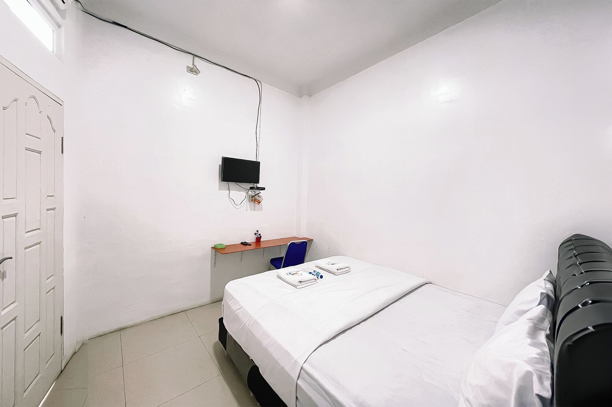 A small, white-walled room with a single bed, a desk with a chair, and a television mounted on the wall.