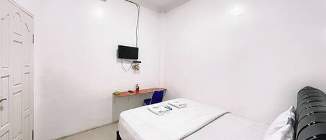 A small, white-walled room with a single bed, a desk with a chair, and a television mounted on the wall.