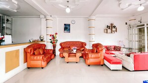 A spacious living room with a red sofa set, a coffee table, and a red and white couch. There is a reception desk with a counter and a shelf with decorative items. - Agave Hotel By Helocus (Pematang Pane)