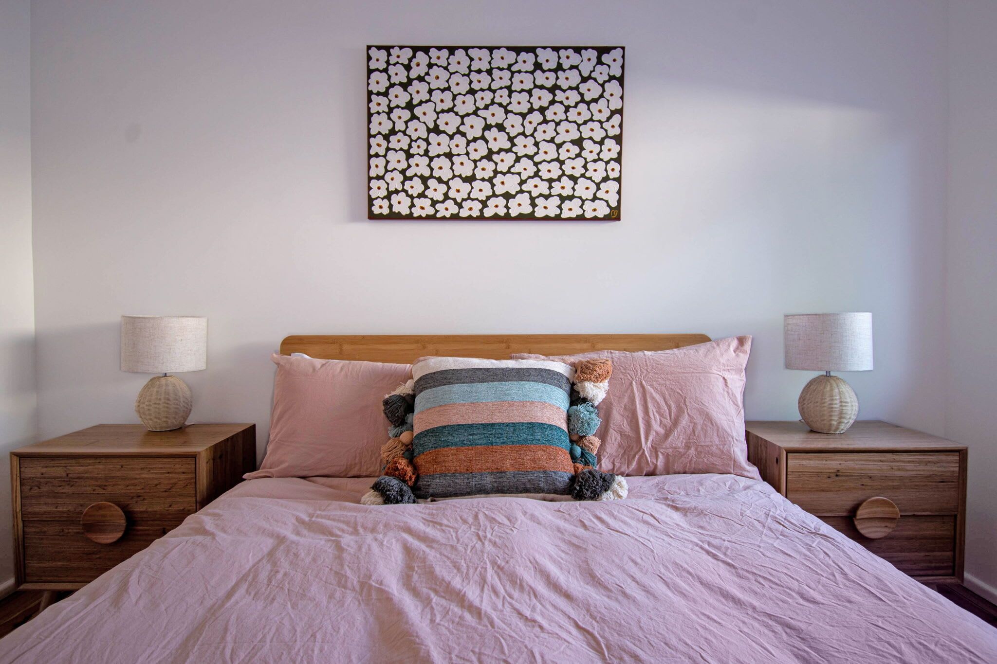 A bedroom with a wooden headboard, a pink bedspread, two bedside tables with lamps, and a wall art piece above the bed.