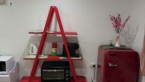 Une petite pièce dans laquelle se trouve une étagère rouge abritant une bouilloire, un mixeur et un four grille-pain. Un vieux réfrigérateur rouge se trouve à droite, et un lit avec une serviette bleue est visible au premier plan.