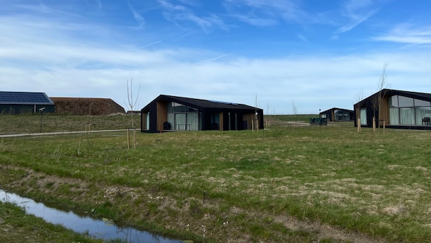 A modern building with large windows and a flat roof, situated in a grassy field with a small water body in the foreground. - Holiday Home in Wissenkerke With Garden (Wissenkerke)