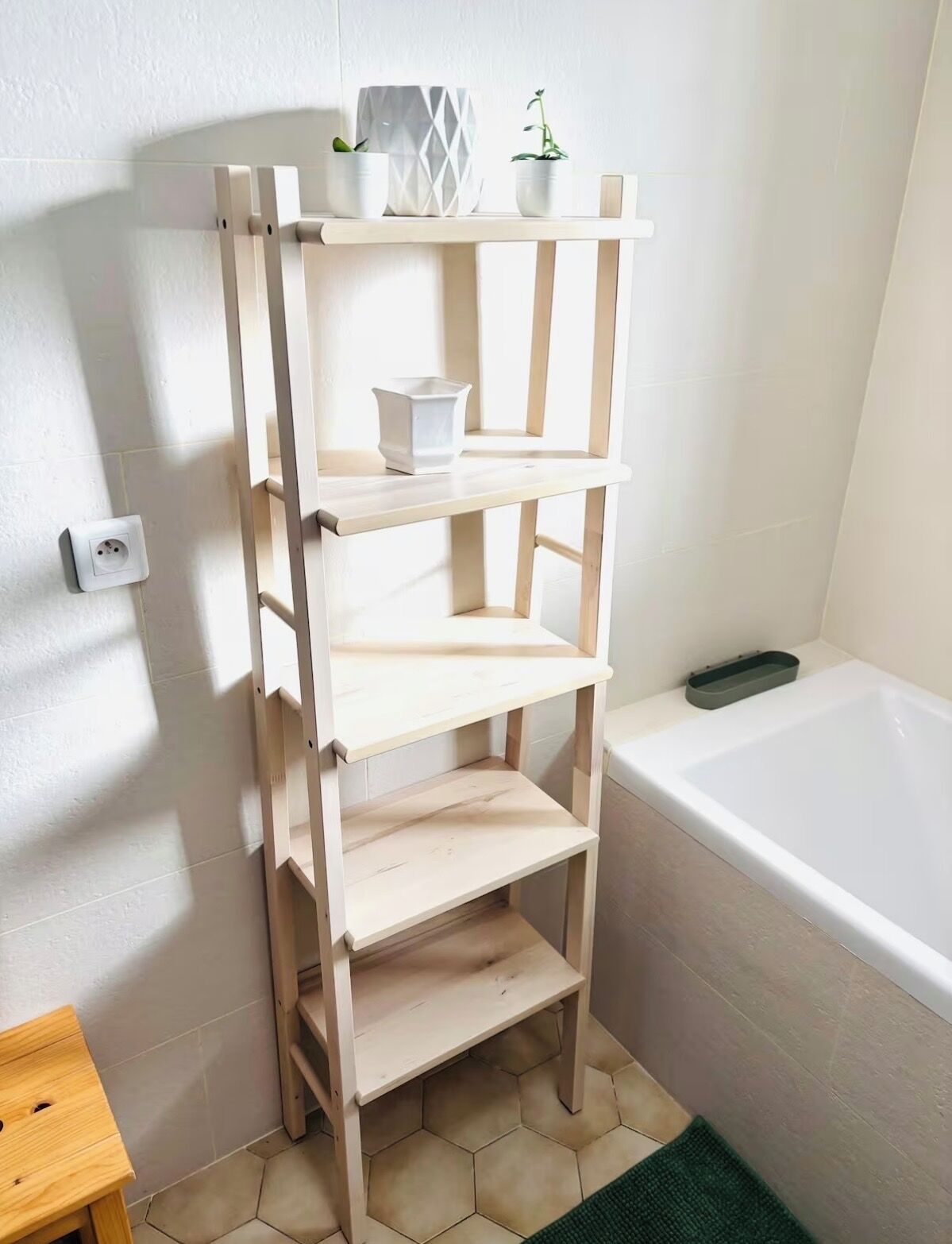 A bathroom with a wooden shelf unit, a white bathtub, and a small wooden stool.