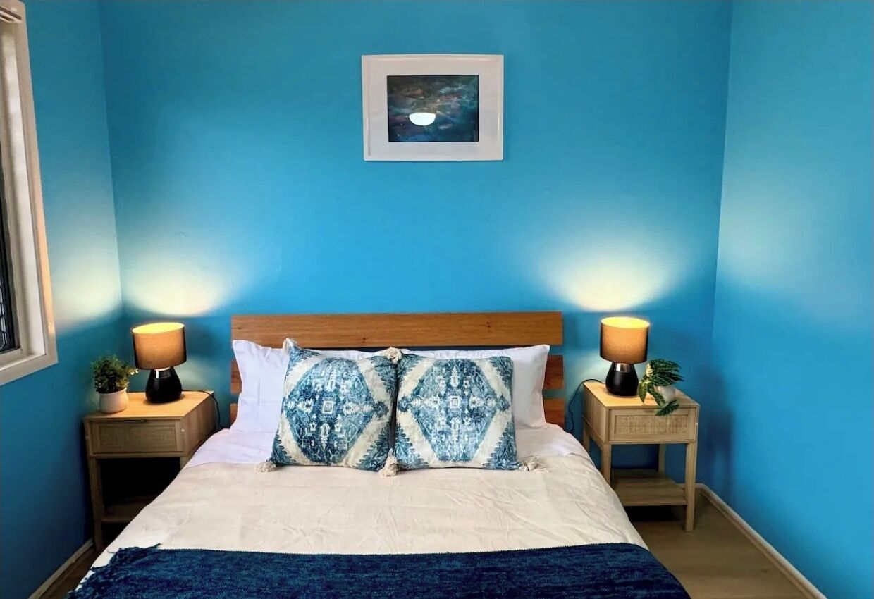 A bedroom with a bed, two bedside tables with lamps, a framed picture on the wall, and a blue wall.